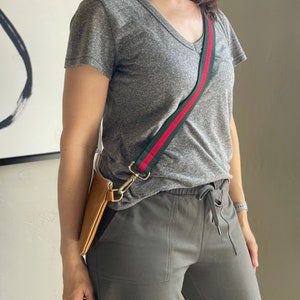 May include: A gray t-shirt with a red and green striped strap. The strap is attached to a brown leather bag. The person is wearing gray pants.