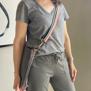 May include: A gray t-shirt with a v-neck and a rainbow striped shoulder strap. The strap is attached to a brown canvas bag. The person is wearing gray sweatpants with a drawstring.