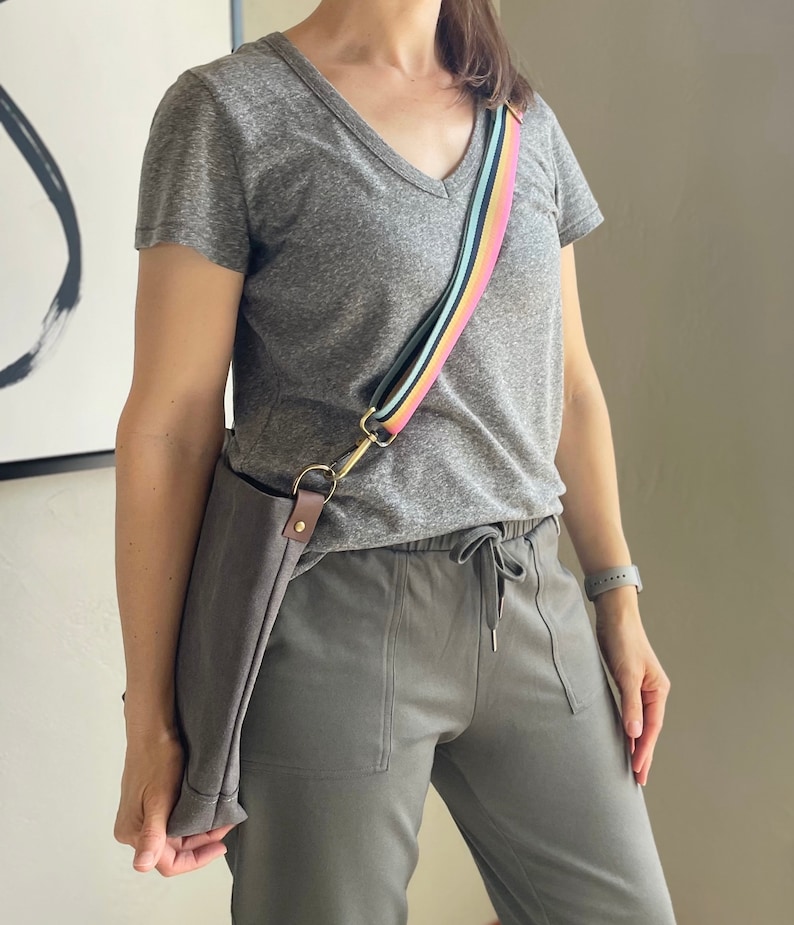 May include: A gray t-shirt with a v-neck and a rainbow striped shoulder strap. The strap is attached to a brown canvas bag. The person is wearing gray sweatpants with a drawstring.