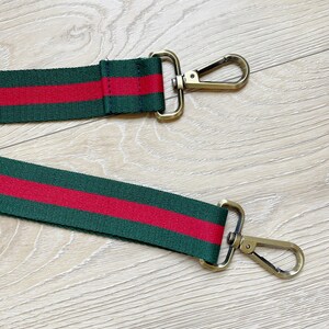 May include: A green and red striped fabric strap with gold metal clasps. The strap is designed to be used as a replacement strap for a bag.