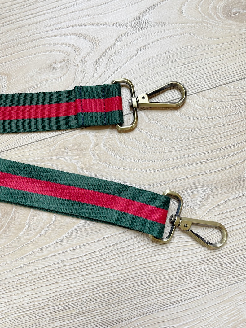 May include: A green and red striped fabric strap with gold metal clasps. The strap is designed to be used as a replacement strap for a bag.