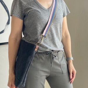 May include: A gray t-shirt with a v-neck and a multi-colored striped strap attached to a black canvas bag. The bag is worn over the shoulder and the strap is attached to a gold clasp.