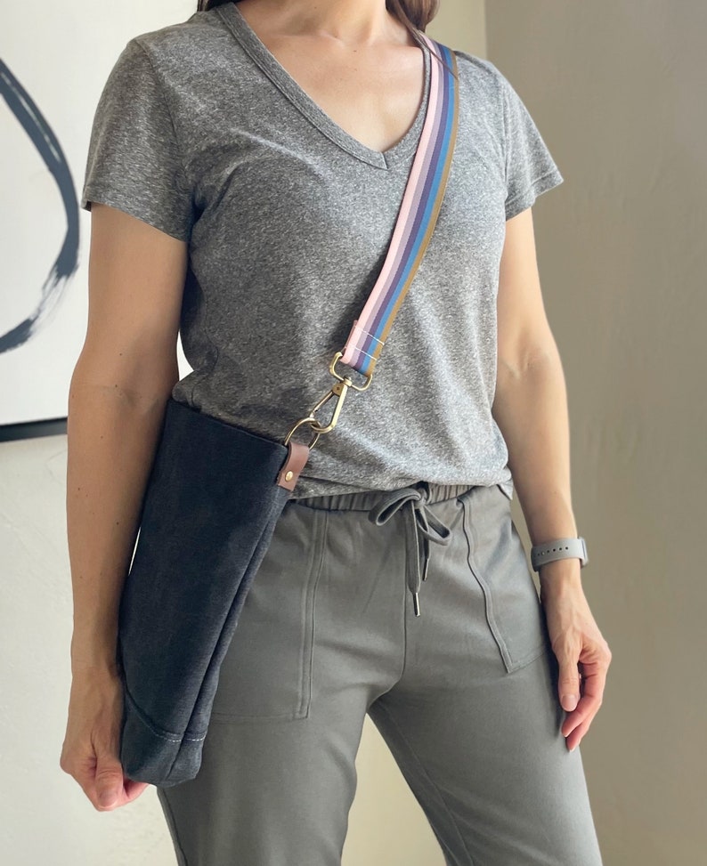 May include: A gray t-shirt with a v-neck and a multi-colored striped strap attached to a black canvas bag. The bag is worn over the shoulder and the strap is attached to a gold clasp.