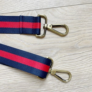 May include: A navy blue and red striped strap with gold metal clasps. The strap is made of woven fabric and has a red stripe running down the center.