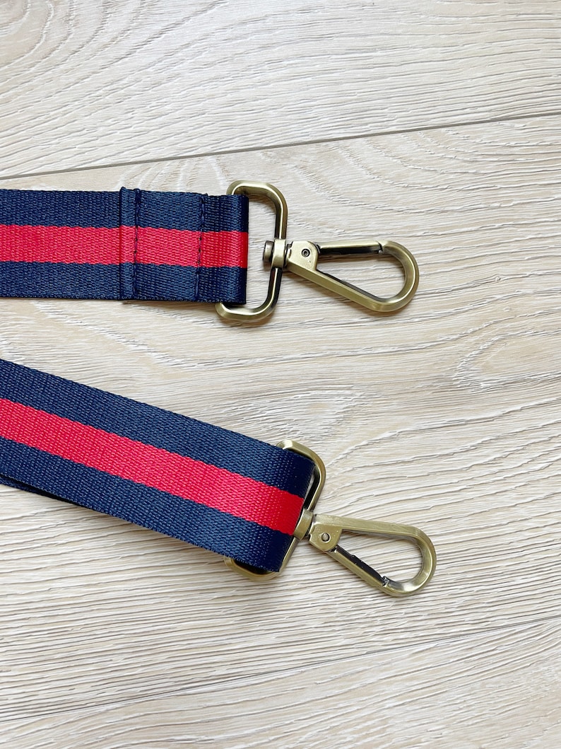May include: A navy blue and red striped strap with gold metal clasps. The strap is made of woven fabric and has a red stripe running down the center.