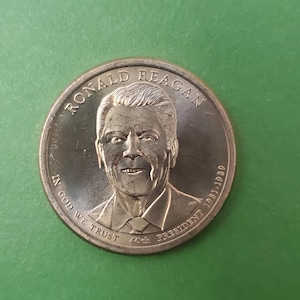 May include: A bronze coin featuring a portrait of Ronald Reagan. The coin is inscribed with "RONALD REAGAN", "IN GOD WE TRUST", "40th PRESIDENT 1981-1989". The coin has a detailed relief of Reagan's face and hair.