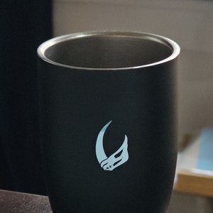 Puede incluir: Una taza de viaje de acero inoxidable negra con un logotipo blanco de una luna creciente y una cabeza de animal estilizada.