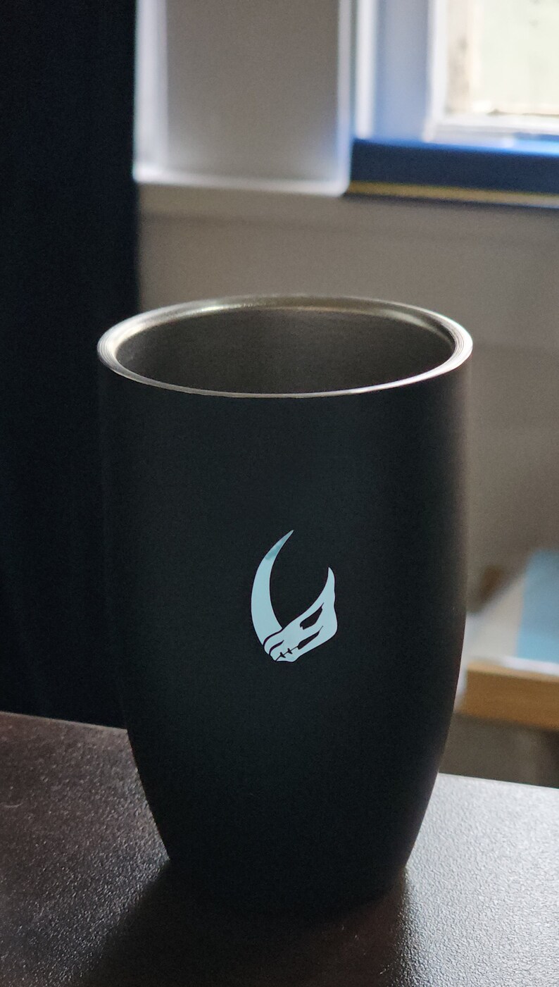 Puede incluir: Una taza de viaje de acero inoxidable negra con un logotipo blanco de una luna creciente y una cabeza de animal estilizada.