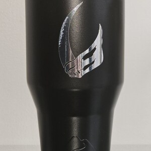Puede incluir: Un vaso de acero inoxidable negro con un s&iacute;mbolo mandaloriano plateado en la parte delantera. El vaso tiene una tapa plateada y una base plateada.