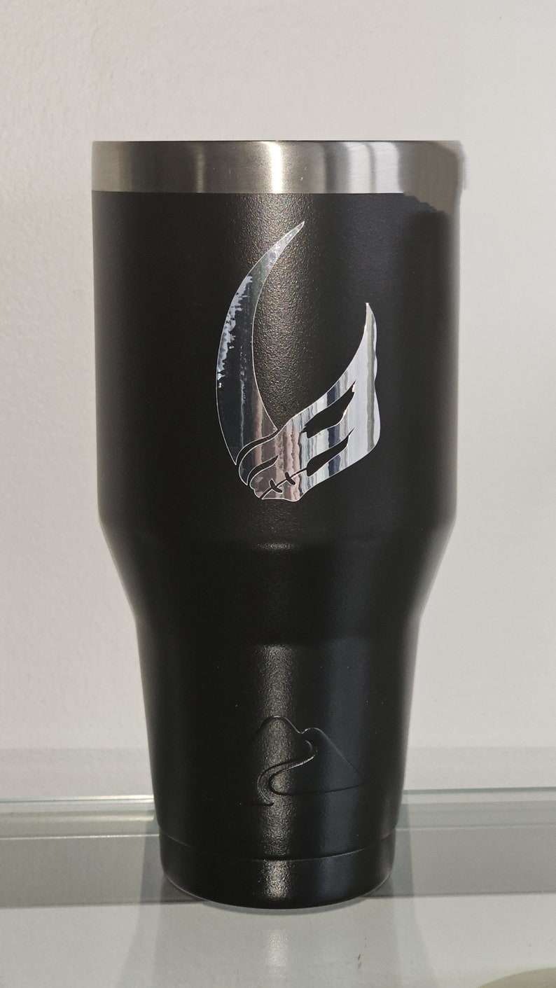 Puede incluir: Un vaso de acero inoxidable negro con un s&iacute;mbolo mandaloriano plateado en la parte delantera. El vaso tiene una tapa plateada y una base plateada.