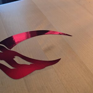 Puede incluir: Una pegatina met&aacute;lica roja de una luna creciente estilizada con una cara dentro.