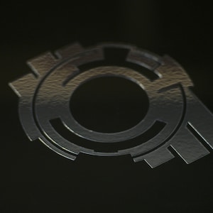 May include: A silver-colored metal logo with a circular design and a central hole. The logo has a textured surface and a three-dimensional appearance.