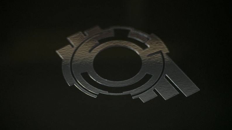 May include: A silver-colored metal logo with a circular design and a central hole. The logo has a textured surface and a three-dimensional appearance.