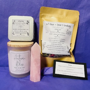 May include: A gift set with a pink rose quartz crystal, a soy wax candle labeled "Autumn Bliss", a tin with the text "a big hug", a brown paper bag with the text "self love + heart healing summer pot", and a black card with the text "Honey".