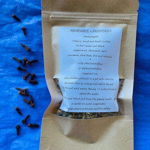 May include: A brown paper pouch with a white label that reads "ABUNDANCE + PROSPERITY simmerpot -organic, hand and heart crafted herbal simmer pot blend- peppermint, chamomile, basil, cinnamon, clove buds, love and intention esty: plantseedsoflove ig: @plantseedsoflove suggested use: place herbal contents in a pot with intention. optional: slice an orange and add to the pot. fill the pot with water, leaving -2 inches of space above the water. wer your blend and enjoy the yummy smells.... in water as water evaporates. ways practice kitchen and fire safety-"