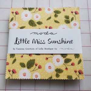 May include: A stack of fabric squares with a yellow background and a floral daisy pattern. The fabric is wrapped with a white label that reads "Little Miss Sunshine" by Vanessa Goertzen of Lella Boutique for moda.