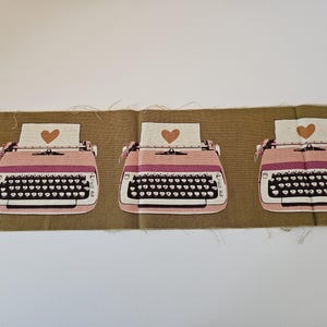 May include: A rectangular piece of brown fabric featuring a repeating pattern of three pink typewriters. Each typewriter has a white sheet of paper inserted into the carriage, with a brown heart printed on each sheet of paper.
