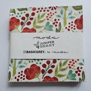 May include: A square stack of fabric squares featuring a cream background with a botanical print of red berries, red flowers, and green leaves. A white paper band wraps around the center with the text moda JUNIPER BERRY BASICGREY for moda.
