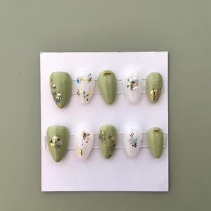 May include: A set of ten almond-shaped false nails. The nails are a mix of solid olive green and white, decorated with gold and blue foil accents. The nails are arranged on a white card against a green background.