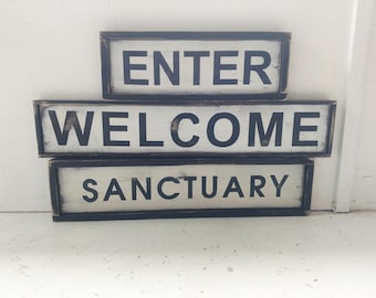 May include: Three wooden signs stacked on top of each other. The top sign reads "ENTER" in black letters on a white background. The middle sign reads "WELCOME" in black letters on a white background. The bottom sign reads "SANCTUARY" in black letters on a white background.