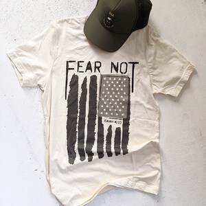 May include: A white t-shirt with a black and white American flag design and the text "FEAR NOT" above the flag. The text "ISAIAH 41:10" is below the flag. A green baseball cap with a USA logo is on top of the shirt.