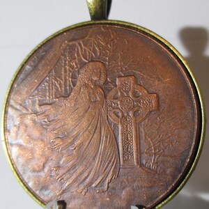 Puede incluir: Un colgante de color cobre con un relieve detallado de una figura encapuchada de pie frente a una cruz celta. El colgante está unido a una cadena de color dorado.