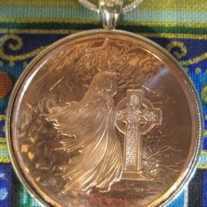 May include: A copper-colored pendant necklace featuring a detailed image of a woman standing next to a Celtic cross. The woman is wearing a flowing robe and the cross is intricately designed with knots and patterns.