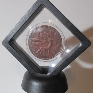Puede incluir: Una moneda de color cobre con un diseño de dragón en una vitrina negra con una cubierta de plástico transparente. La moneda está en un soporte negro.