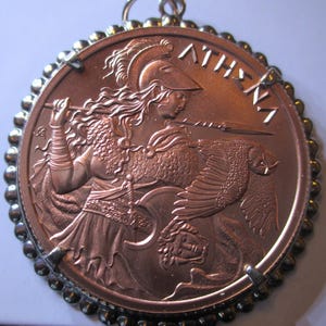 May include: A copper-colored pendant featuring a detailed relief of Athena, the Greek goddess of wisdom and warfare, holding a spear. The word "ATHENA" is inscribed above the figure. The pendant is framed by a beaded border and includes a loop for hanging.