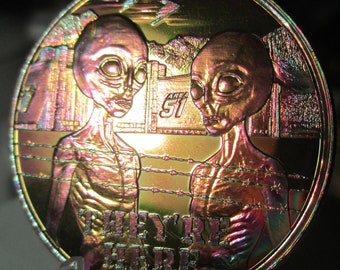 May include: A gold-coloured coin with a holographic finish features two grey alien figures in front of a barbed wire fence and a building with the text "Area 51". The text "They're Here" is printed below the figures.