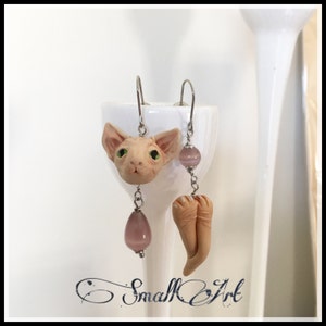 以下が含まれることがあります： ピンクの猫の頭とピンクの猫の足がついたイヤリングと、ピンクの宝石がついたイヤリングのペアです。