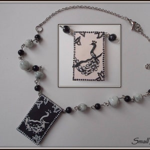 May include: A black and white necklace with a pendant featuring a peacock design. The pendant is made of black and white paint on a rectangular piece of material. The necklace is made of a silver chain and black and white beads.