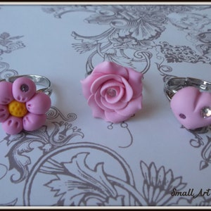 Può includere: Tre anelli a forma di fiore rosa con bande argentate. Gli anelli presentano una rosa rosa, un fiore rosa con un centro giallo e un cuore rosa con un strass.
