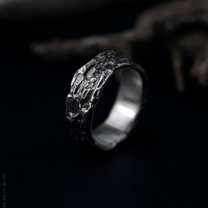 May include: A silver ring with a textured, bark-like design. The ring is thick and has a wide band.