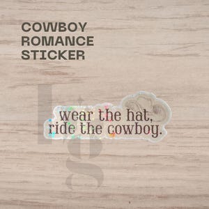 May include: A clear sticker with the text "wear the hat, ride the cowboy." The sticker has a cowboy hat graphic and the words "Cowboy Romance Sticker" in black. The sticker has iridescent glitter accents.
