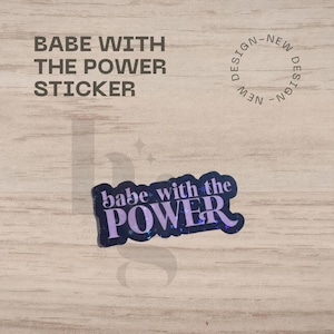 May include: A sticker with the text "babe with the POWER" in purple and white, with a dark blue background. The sticker is on a light brown wooden surface. The words "BABE WITH THE POWER STICKER" are printed above the sticker.