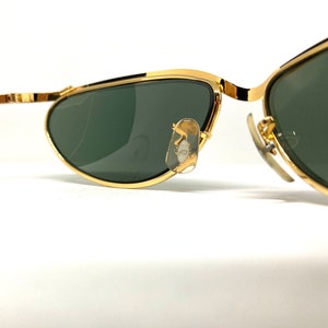 May include: Gold-tone aviator sunglasses with green lenses. The sunglasses have a thin metal frame and are designed with a curved shape.