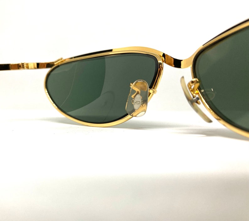 May include: Gold-tone aviator sunglasses with green lenses. The sunglasses have a thin metal frame and are designed with a curved shape.