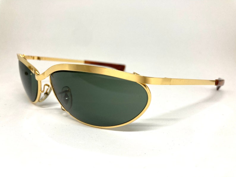 May include: Gold-tone aviator sunglasses with green lenses. The sunglasses have a thin metal frame and a curved, oval shape.