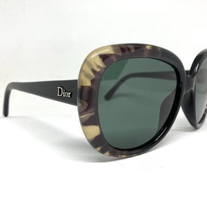 May include: Oversized sunglasses with a black frame and green lenses. The outer frame has a tortoiseshell pattern. The word "Dior" is printed on the left arm of the sunglasses.