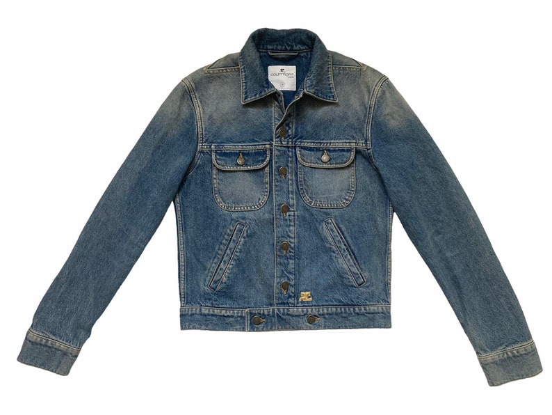 May include: A blue denim jacket with a button closure, two chest pockets, and a small logo patch on the bottom right pocket.