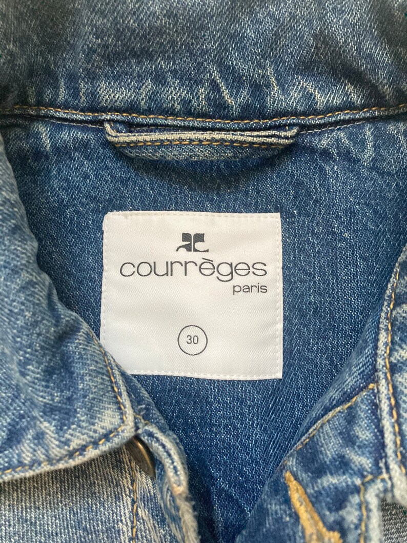 May include: A white label with the text "Courr&egrave;ges Paris" and a size 30 tag attached to a blue denim jacket.