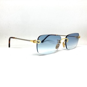 Op de afbeelding: Randloze zonnebril met een blauwe gradiënttint. De monturen zijn goudkleurig met een gevlochten zilverkleurig montuur en schildpadkleurige oortips. De zonnebril staat tegen een witte achtergrond.