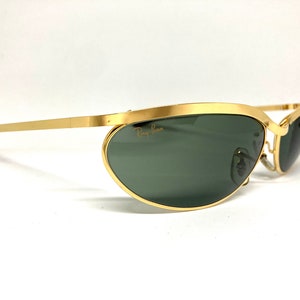 May include: Gold-tone Ray-Ban sunglasses with green lenses and a curved, oval shape. The sunglasses have a thin metal frame and a brown temple.