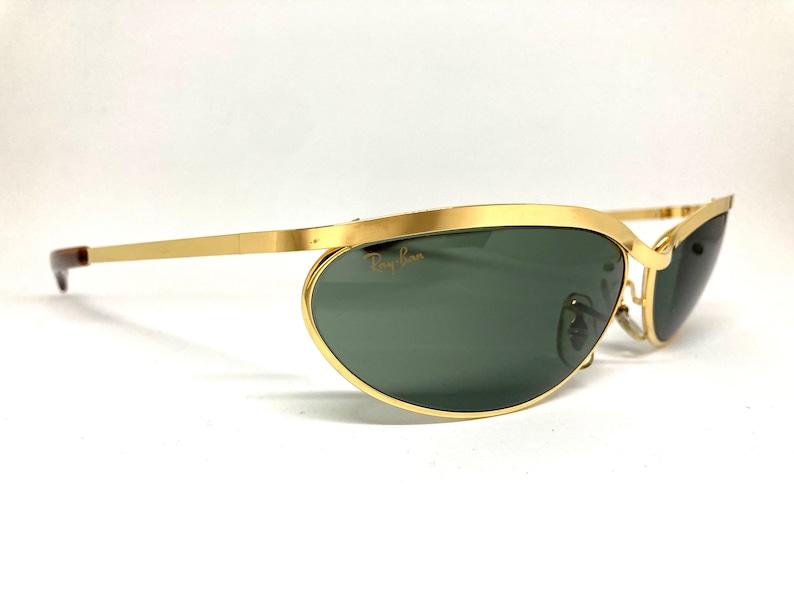 May include: Gold-tone Ray-Ban sunglasses with green lenses and a curved, oval shape. The sunglasses have a thin metal frame and a brown temple.
