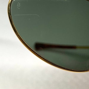 May include: Close-up of a gold-rimmed, round, green-tinted sunglass lens with the word "Ray-Ban" inscribed on the top edge. The lens also has a small "B" inscribed on the left side.