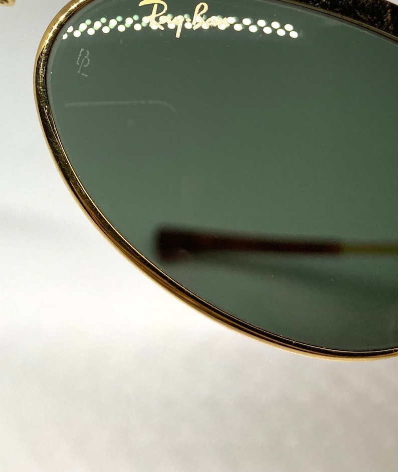 May include: Close-up of a gold-rimmed, round, green-tinted sunglass lens with the word "Ray-Ban" inscribed on the top edge. The lens also has a small "B" inscribed on the left side.