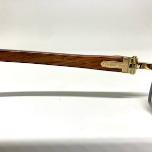 May include: Gold-tone Cartier Paris sunglasses with a wooden frame and blue gradient lenses. The frame is engraved with "Made in France, CE 140 B".
