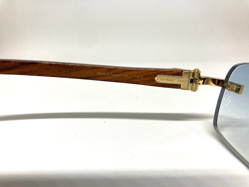 May include: Gold-tone Cartier Paris sunglasses with a wooden frame and blue gradient lenses. The frame is engraved with "Made in France, CE 140 B".