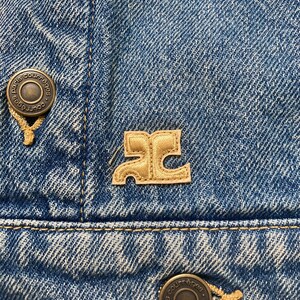 May include: A close-up of a blue denim jacket with two metal buttons that say "Paris Courr&egrave;ges" and a brown leather patch with the letter "C" in a stylized font.